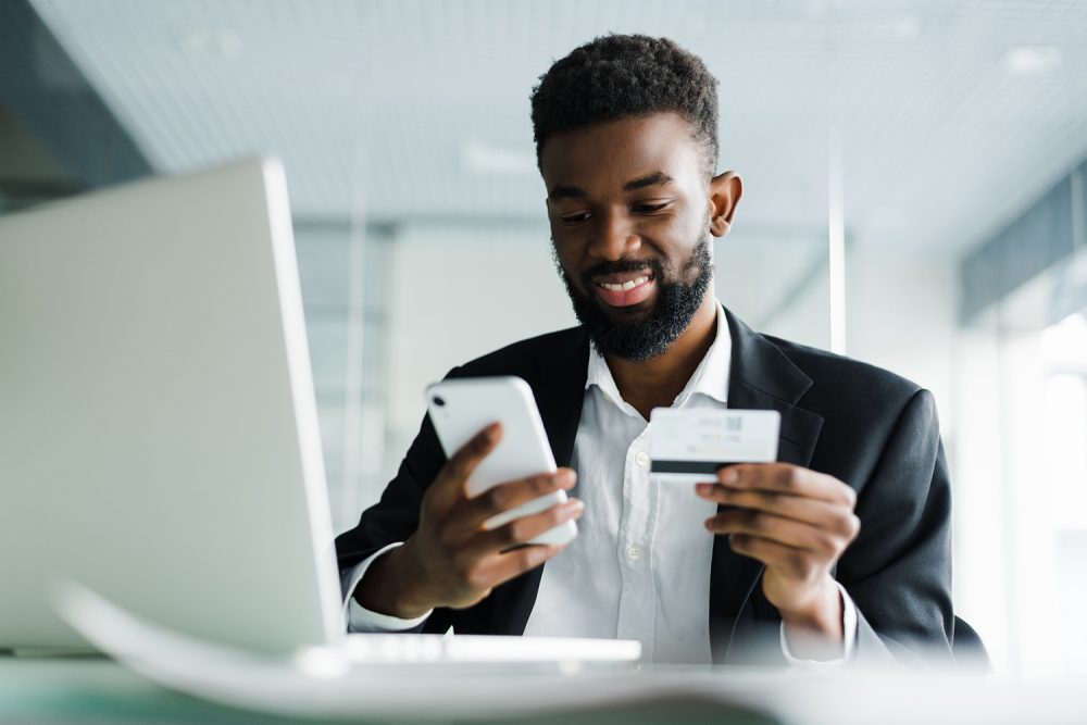 African American man in casual yellow tshirt paying with credit card online while making orders via mobile Internet. Attractive black hipster male making transaction using mobile bank application.